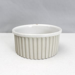 Może przedstawiać: Mały, kremowy ceramiczny ramekin z żłobionym zewnętrzem. Okrągłe naczynie ma lekko teksturowaną krawędź i jest przeznaczone do pieczenia i serwowania pojedynczych porcji jedzenia. Ramekin jest ustawiony na białym tle.