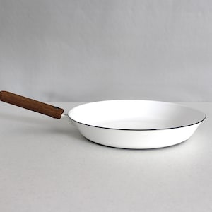 May include: A white frying pan with a dark blue rim and a wooden handle. The pan is round and has a simple, minimalist design. The handle is made of a light brown wood.