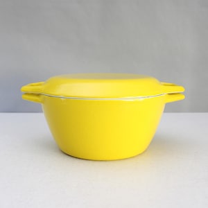 Puede incluir: Una fuente para horno de color amarillo brillante con tapa. El plato tiene forma redondeada y dos asas pequeñas. La tapa encaja perfectamente, con una fina línea blanca que acentúa el borde. El plato está sobre un fondo neutro.