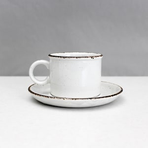 Op de afbeelding: Een witte keramische koffiekop en schotel set. De beker heeft een klein handvat en een gespikkelde afwerking met een donkerbruine rand. De schotel heeft een bijpassende gespikkelde afwerking en een donkerbruine rand. De set staat op een wit oppervlak.