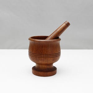 May include: A wooden mortar and pestle set. The mortar is a small, round bowl with horizontal grooves, and the pestle is a cylindrical stick. Both pieces are made of a dark brown wood.