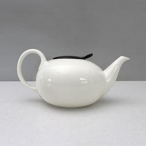 Peut inclure: Une théière en céramique blanche avec un corps arrondi, une anse incurvée et un bec verseur. La théière a un couvercle noir et est posée sur un fond neutre. Cette théière est idéale pour servir le thé.