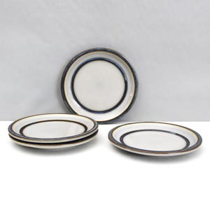 May include: A set of four ceramic plates. The plates have a white center with a dark brown ring and a blue rim. The plates are arranged on a white surface against a light gray background. The plates are likely for serving food.
