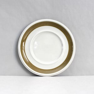 Może przedstawiać: Biały ceramiczny spodek z brązowym paskiem wokół krawędzi i mniejszym brązowym paskiem w pobliżu środka. Spodek jest okrągły i stoi na białej powierzchni.