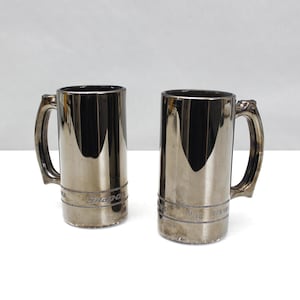 May include: Two silver-colored beer steins with black interiors and handles. The steins have a metallic finish and a band of text near the base. The steins are cylindrical and sit on a white surface.