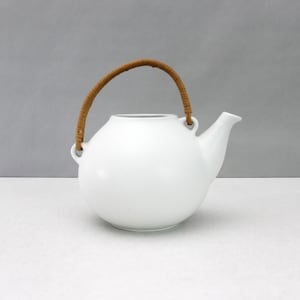May include: A white ceramic teapot with a spherical body and a short, angled spout. The teapot features a curved, brown, textured handle made of wrapped material that attaches to two small loops on the top sides of the white ceramic vessel.