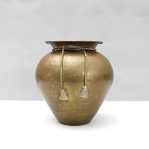 Peut inclure: Un vase en laiton martelé, de forme arrondie et à bord évasé. Il est orné d'un détail de corde autour du col, avec deux glands décoratifs. Le vase a une teinte dorée chaude et est idéal pour la décoration.