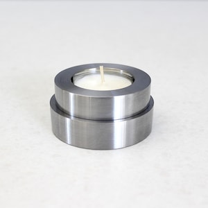 Op de afbeelding: Een moderne kaarsenhouder van geborsteld metaal met een kleine witte kaars. De kaarsenhouder bestaat uit twee gestapelde, cirkelvormige metalen onderdelen. De kaars is ongeveer 2,5 cm in diameter.