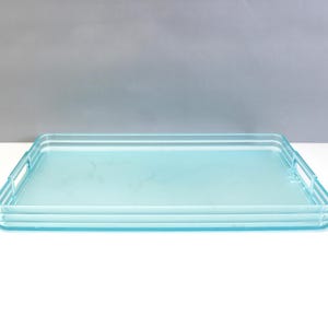 May include: A rectangular, light blue acrylic serving tray with a smooth, translucent finish. The tray features integrated handles on both short sides and a tiered, layered edge design that creates a subtle ribbed effect along the perimeter.