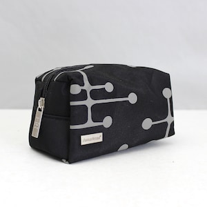 Könnte beinhalten: Schwarze, rechteckige Kosmetiktasche mit silbernem Reißverschluss und grauem geometrischem Muster. Die Tasche hat einen kleinen silbernen Anhänger am Reißverschluss und ein silbernes Etikett mit der Aufschrift "Amadea".