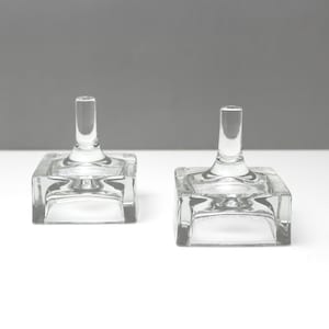 Puede incluir: Dos candelabros de vidrio transparente. Cada uno tiene una base cuadrada y un tallo cilíndrico. El vidrio es transparente, permitiendo el paso de la luz. Los candelabros están colocados sobre una superficie blanca sobre un fondo gris.