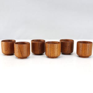 May include: Six small, handcrafted wooden cups with a natural wood grain finish. The cups are cylindrical with a slightly flared rim and a rounded base. The wood has a warm, brown tone, and the cups are arranged in a row.