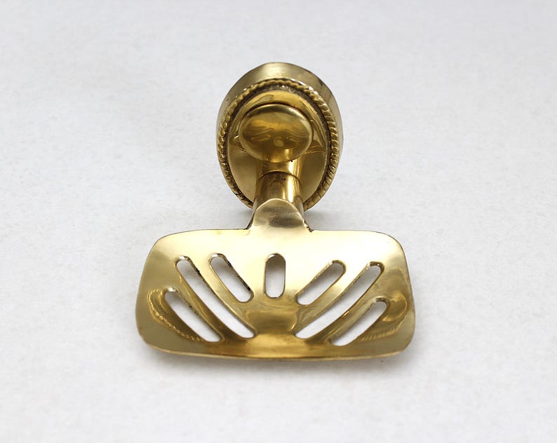 May include: A brass soap dish with a decorative, oval-shaped backplate and a slotted tray. The dish has a shiny, golden finish and is designed for wall mounting. The tray has a series of curved slots for drainage.
