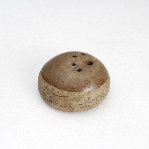 May include: A small, round ceramic salt or pepper shaker with a smooth, glossy brown glazed top and a textured, speckled beige base. The top surface features four small, evenly spaced holes for dispensing contents, creating a simple, functional design.