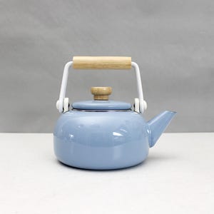 Puede incluir: Una tetera de esmalte azul claro con asa y pomo de tapa de madera. El asa es blanca con un agarre de madera clara. La tetera tiene un pico para verter y está sobre un fondo neutro.