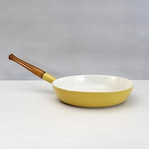 May include: A yellow frying pan with a white interior and a wooden handle. The pan has a rounded shape and is designed for cooking. The handle is a light brown color and is attached to the pan with a yellow base.