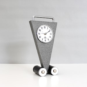 Puede incluir: Un reloj gris y plateado con una esfera blanca y agujas negras. El reloj está en un soporte con dos ruedas negras. El reloj está sobre una superficie blanca.