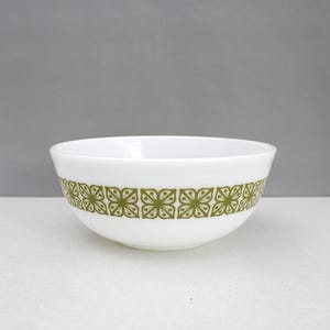 May include: A white glass mixing bowl featuring a repeating olive green floral pattern around the exterior. The bowl has a smooth, rounded shape with a wide opening and a clean, white interior surface, showcasing a classic mid-century design.