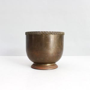 Puede incluir: Un cuenco redondo de latón con una pátina lisa y desgastada y un acabado metálico oscuro. El borde superior presenta una banda decorativa con un patrón floral repetitivo, y el cuenco descansa sobre una base circular ligeramente elevada con un borde cobrizo.