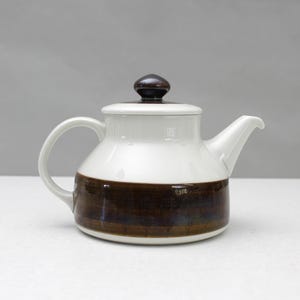 Może przedstawiać: Ceramiczny czajnik z białą górną częścią i brązowym pasem wokół podstawy. Czajnik ma pasującą pokrywkę z ciemnobrązowym guzikiem i zakrzywionym uchwytem. Dzióbek jest biały i ustawiony pod kątem.