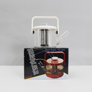 May include: A clear glass Bistro teapot with a white handle and lid, sitting atop a black box. The teapot has a stainless steel infuser and a spout. The box features a diagram of the teapot and the text "Bistro Teapot / 0.5 Liter".