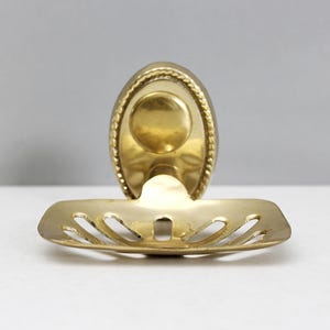 May include: A vintage brass soap dish with an oval back and a slotted tray. The dish has a decorative rope-like border and a circular indentation for the soap. The brass has a warm, golden hue.