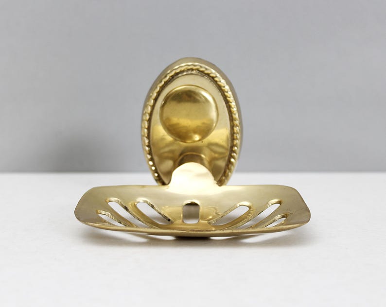 May include: A vintage brass soap dish with an oval back and a slotted tray. The dish has a decorative rope-like border and a circular indentation for the soap. The brass has a warm, golden hue.