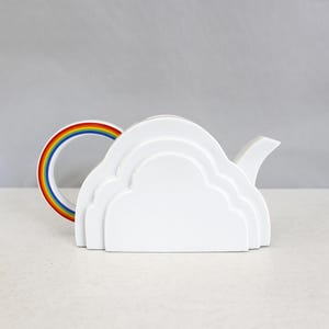 Puede incluir: Tetera de cerámica blanca con forma de nube, con un asa de arcoíris. La tetera tiene un pico vertedor y un diseño de nube en capas en el cuerpo. El asa presenta los colores del arcoíris.