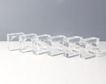 Op de afbeelding: Set van zes transparante acryl servetringen. De ringen zijn vierkant met een ronde opening in het midden.