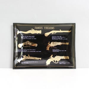 May include: A black rectangular plaque featuring gold illustrations of antique firearms. The plaque is labelled "FAMOUS FIREARMS" and includes text detailing each gun's origin and type. The plaque is set against a white background.