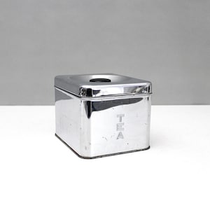 May include: A square, polished chrome tea canister with a removable lid featuring a small black circular knob. The front side of the metallic container is embossed with the word TEA in vertical capital letters, reflecting light on its surface.