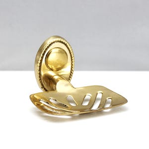 May include: A vintage brass soap dish with an oval backplate and a leaf-shaped holder. The backplate has a rope-like border and a central circular detail. The holder features a series of elongated cutouts.