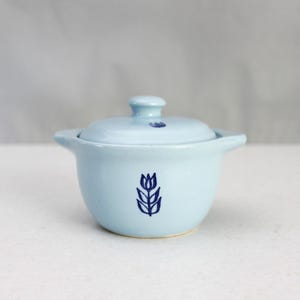 Könnte beinhalten: Hellblaue Keramiktopf mit Deckel und zwei kleinen Griffen. Der Topf hat ein dunkelblaues Tulpenmuster auf der Vorderseite und einen kleinen Knopf auf dem Deckel. Der Topf steht auf einer weißen Oberfläche.