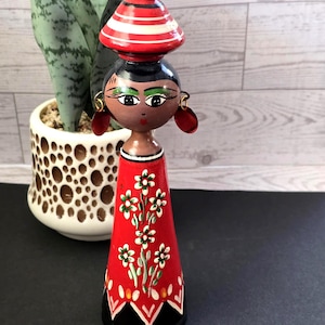 May include: A hand-painted wooden figurine of a woman with a red dress adorned with white flowers. The figurine has a black base, a brown face with painted features, and a red and white striped hat. Red earrings complete the look.