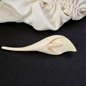 May include: Ivory-colored calla lily brooch with a detailed stamen, set against a dark background. A matching ivory fabric, possibly a scarf or headband, is visible in the background. The brooch is a delicate floral accessory.