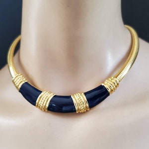 May include: A gold-toned necklace with a navy blue enamel center. The necklace has a snake chain design with gold accents and a curved, geometric shape. The necklace is displayed on a mannequin.