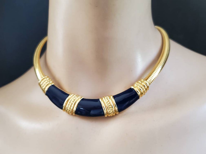 May include: A gold-toned necklace with a navy blue enamel center. The necklace has a snake chain design with gold accents and a curved, geometric shape. The necklace is displayed on a mannequin.