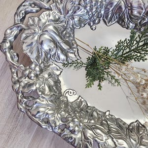 May include: Silver-toned metal tray with a decorative grape and leaf design around the edges. The tray has a rectangular shape with rounded corners. The tray is decorated with a bunch of grapes and leaves. The tray is empty.