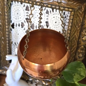 May include: A polished copper bowl with a smooth, rounded interior and exterior, suspended by three thin metal chains that connect at a single top point. The bowl has a warm, metallic finish and a simple, open-top design.