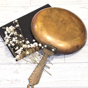 May include: A vintage brass pan with a long handle and a hammered texture. The pan is oval-shaped and has a golden hue. A black book and white flowers are in the background.