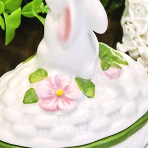 May include: A white ceramic bunny figurine atop a white basket-weave container with a green rim. The bunny has pink inner ears and is surrounded by pink flowers and green leaves. A charming Easter-themed decorative piece.