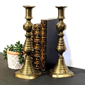 May include: Two antique brass candlesticks with geometric designs. The candlesticks have a wide base and a detailed stem, with a top designed to hold a candle. A small potted plant and a stack of books are in the background.