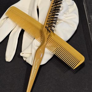 May include: Mustard-colored hair comb and brush set with a white glove on a black surface. The brush has black bristles and a curved handle. The comb is straight with fine teeth. The brush handle reads "The Little Wonder".