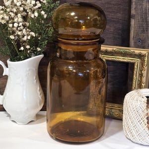 May include: A vintage amber glass apothecary jar with a round stopper. The jar is cylindrical with a wide base and a narrow neck. The jar is empty and sits on a white surface. A white ceramic pitcher with flowers is to the left.