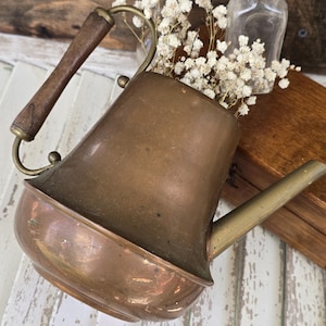 May include: A vintage copper watering can with a wooden handle and brass accents. The can is filled with white flowers and sits next to a small glass bottle. The copper has a warm, aged patina.