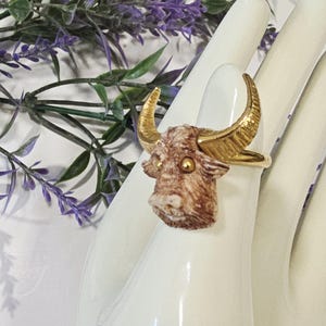 May include: A gold-toned ring featuring a detailed bull head with textured brown and white fur and gold horns. The ring is set against a white background with green and purple foliage.