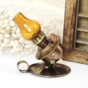 May include: A miniature antique oil lamp with a golden glass shade and a brass base. The lamp is tilted, showcasing its intricate details, including a small handle and a decorative rim. The lamp sits on a round base.