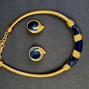 May include: A gold-toned necklace and matching earrings set. The necklace features a flexible, ribbed design with a section of dark blue enamel and gold bands. The earrings have a matching dark blue enamel swirl design. The jewelry set is displayed on a dark gray background.