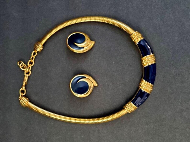 May include: A gold-toned necklace and matching earrings set. The necklace features a flexible, ribbed design with a section of dark blue enamel and gold bands. The earrings have a matching dark blue enamel swirl design. The jewelry set is displayed on a dark gray background.