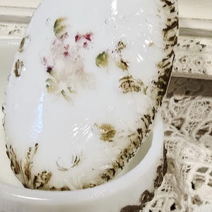 May include: White oval trinket box with a lid. The lid features a hand-painted floral design in pink, green, and brown. The edges of the lid and base have a brown, distressed trim. The box is sitting on a white lace surface.